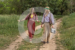 Couple of young people in hippie style on a country road. The girl is dancing, the guy is carrying a retro cassette recorder