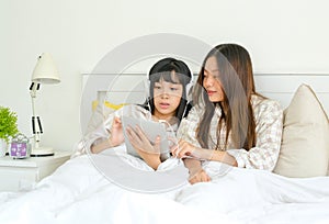 Couple of young sisters talk together and look to the tablet on bed in bedroom with morning light