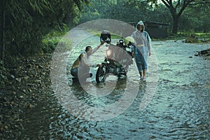 couples of asian biker wearing plastic rain clothes maintenance small enduro motorcycle in shallow creek among rain falling