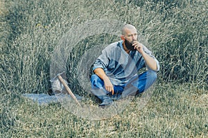 Courageous man with  beard resting in the grass in field. Pensive gaze. Next to him is  ax and  warrior`s helmet