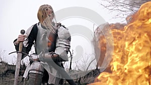 Courageous woman warrior in armor sits leaning on sword against fire