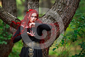 Courageous young lady with long red hair in image of fabulous historical character of witch and priestess in a mystical forest