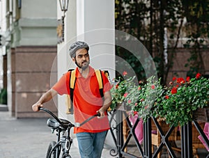 Courier with beard, protective helmet and yellow bag, walking the street with bicycle and looking for client address