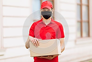 A courier employee in uniform and a protective face mask holds an empty cardboard box outdoors