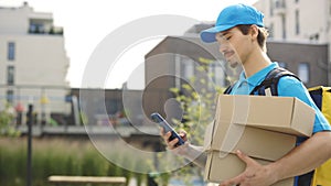 Courier Going Along Building Holding the Paper Boxes and Mobile Phone in Hand. Delivery Man Looking for Delivery Address