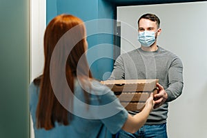 Courier male wearing medical face mask delivering boxes with hot pizza to unrecognizable female customer on doorstep at