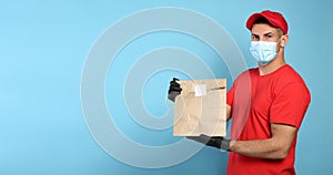 Courier in medical mask holding paper bag with takeaway food on light blue background, space for text. Delivery service during