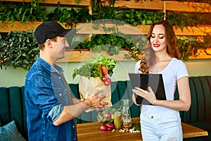 Courier service worker delivering fresh food to a happy woman client signing some documents on the kitchen at home. Online grocery