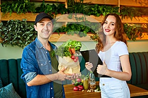 Courier service worker delivering fresh food to a happy woman client signing some documents on the kitchen at home. Online grocery