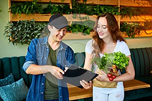 Courier service worker delivering fresh food to a happy woman client signing some documents on the kitchen at home. Online grocery