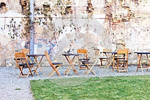 Courtyard outdoor cafe area with wooden tables and chair, green grass lawn and ruined shbby brick wall on the background. Open air