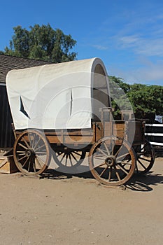 Covered wagon with dirt foreground and green tree, blue sky background with partial fence and building photo