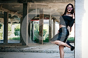A coveted woman or girl stands in the middle of an abandoned building in a leather skirt top and sunglasses, brunette