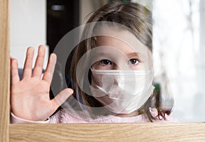 COVID-19 coronavirus concept, little girl in face mask looking through window at home or clinic. Portrait of sad kid during