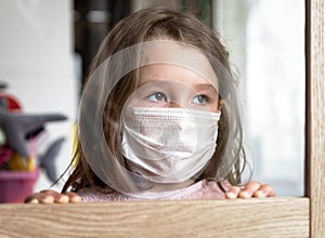 COVID-19 coronavirus concept, little girl in face mask looking through window at home or clinic. Portrait of sad kid during