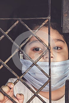 COVID-19 coronavirus concept, little girl in medical mask looking through window at home. Sad kid during quarantine due to COVID