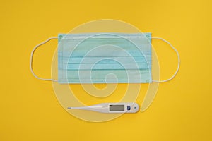 COVID-19 disposable surgical face mask and digital thermometer on yellow background