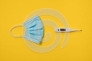 COVID-19 disposable surgical face mask and digital thermometer on yellow background