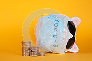 Covid-19 financial crisis. Coronavirus crisis, white piggy bank with face mask stuffed with coin on yellow background