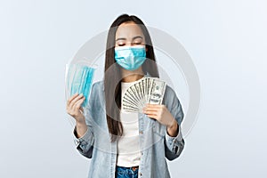 Covid-19 pandemic, coronavirus expences and finance concept. Satisfied greedy asian girl in medical mask hugging money