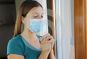 COVID-19 Pandemic Coronavirus Woman Praying Wearing Mask Against Virus SARS-CoV-2. Girl with closed eyes praying near the window