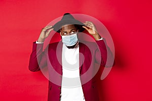 Covid-19, quarantine and holidays concept. Handsome and stylish african american man in face mask, put hat on head and
