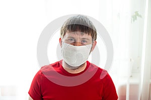 COVID-19. Self-isolation. Young caucasian man in a white mask and red t-shirt in the kitchen during a pandemic. Coronavirus.