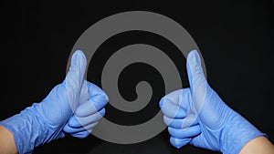 COVID-19 Two hands covered in blue surgical gloves, raising thumbs up, on black background