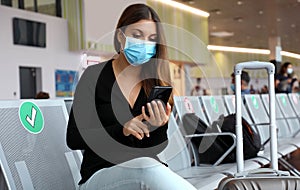 COVID-19 Young tourist woman with surgical mask using phone and sitting respecting social distancing at the airport