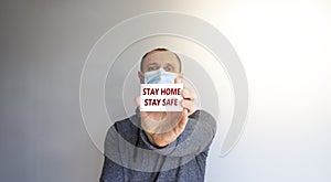 COVID-19 Pandemic symbol. White card with words `Stay home stay safe`. A young strong man in a grey wear and medical mask.