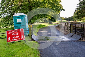 A Covid-19 social distancing warning sign and a plastic portable toilet next to a cattle grid on a public footpath at an outdoor
