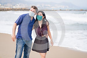 Covid19 beach holidays - young beautiful and happy mixed ethnicity couple of Asian woman and Caucasian man in face mask walking