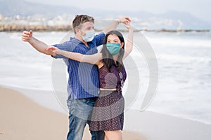 Covid19 beach holidays - young beautiful and happy mixed ethnicity couple of Asian woman and Caucasian man in face mask walking