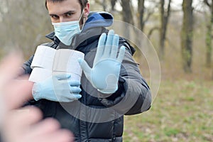Covidiot concept. Young man in protective mask holds many rolls of toilet paper and shows stop gesture outdoors in spring wood