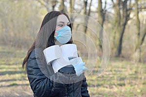 Covidiot concept. Young woman in protective mask holds many rolls of toilet paper outdoors in spring wood