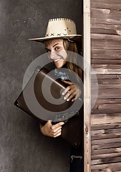 Cowboy girl or pretty woman in stylish hat and blue plaid shirt holding gun and old suitcase