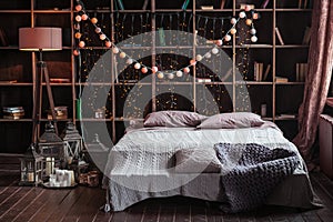 Coziness, comfort, interior and holidays concept - cozy bedroom with bed and garland lights at home. A rack with books