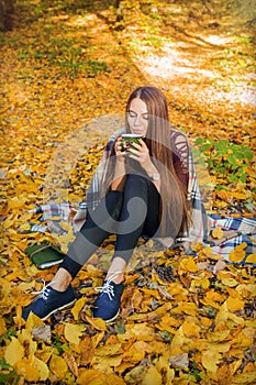 Cozy autumn model photo in yellow leaves. Girl sitting in the autumn forest, in plaid and drinks cofee.