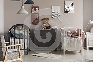 Cozy bedroom interior for twins with white and gray baby beds, a chest of drawers and a fluffy rug. Real photo