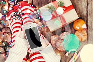 Cozy Christmas at home. Female hands holding mobile phone. Women`s feet in Christmas stockings, big present on bokeh background