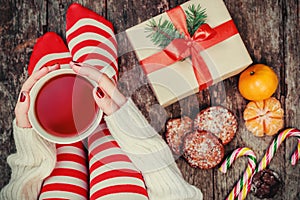 Cozy Christmas at home. Women`s feet in Christmas stockings, big present, cup of tea, candy and Cookies Chocolate on bokeh light b