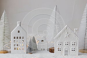 Cozy christmas miniature village. Stylish little ceramic houses and trees on snow blanket with golden lights on white background.