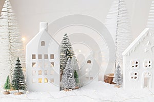 Cozy christmas miniature village. Stylish little ceramic houses and trees on snow blanket with golden lights on white background.