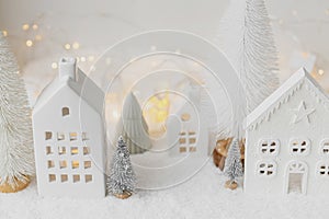 Cozy christmas miniature village. Stylish little ceramic houses and trees on snow blanket with golden lights on white background.