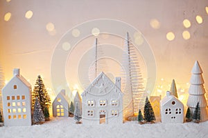 Cozy christmas white miniature village. Stylish little ceramic houses and trees on snow blanket with golden lights in evening.