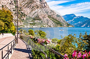 Cozy city street of Limone Sul Garda with paving stone sidewalk, blooming flowers on a metal railings, growing trees with amazing