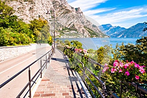 Cozy city street of Limone Sul Garda with paving stone sidewalk, blooming flowers on a metal railings, growing trees with amazing