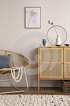 Cozy composition of living room interior with mock up poster frame, rattan comode, stylish armchair, blue pillow, vase with dried