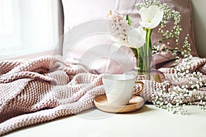 Cozy Easter, spring still life scene. Cup of coffee, pink knitted plaid and floral bouquet in vase on windowsill