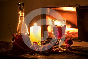 Cozy evening still life by . A glass of red wine, an old decanter, green leaves and dried flowers against a burning fire. Dark pho